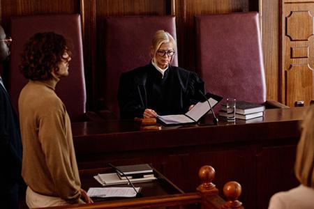 "A judge reviewing a document in a courtroom surrounded by attorneys and other people."